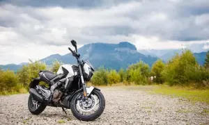 Biało-czarny motocykl Bajaj 400 na kamienistym podjeździe, w tle góry i chmury.