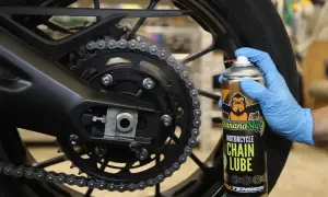 Ręka w niebieskiej rękawiczce aplikuje smar do łańcucha motocyklowego z puszki Banana Slip.