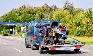 Jak przewieźć motocykl na zwykłej przyczepie? Dwa motocykle bezpiecznie zamocowane pasami na przyczepce za niebieskim vanem.