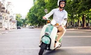 Mężczyzna w kasku i okularach jedzie stylowym, zielono-białym motorowerem na dowód osobisty po brukowanej ulicy.