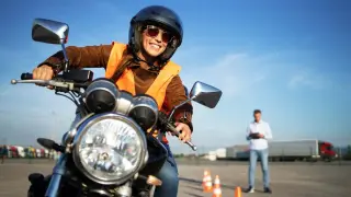 Uśmiechnięta kursantka w kamizelce i kasku na motocyklu, gotowa do zdobycia prawa jazdy na motocykl. Instruktor obserwuje w tle.