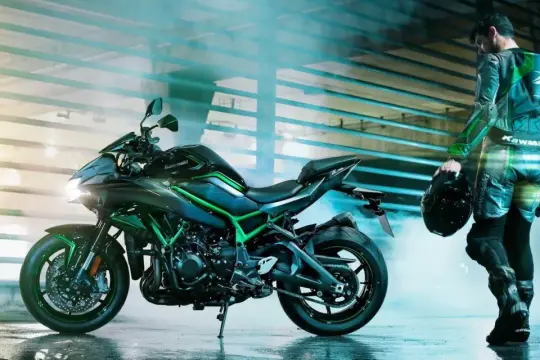 Motocyklista w kombinezonie Kawasaki z kaskiem w dłoni stoi obok czarnego motocykla H2R z zielonymi akcentami.