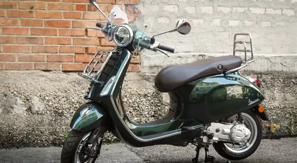 Elegancki, ciemnozielony skuter Piaggio z brązowym siedzeniem i chromowanymi dodatkami stoi na tle ceglanej ściany.