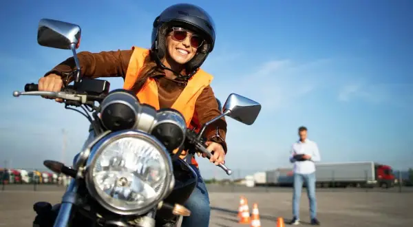 Uśmiechnięta kursantka w kamizelce i kasku na motocyklu, gotowa do zdobycia prawa jazdy na motocykl. Instruktor obserwuje w tle.