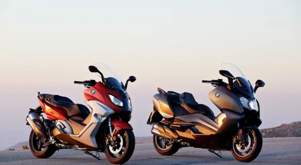 Dwa skutery BMW C650 Sport w kolorach czerwonym i brązowym stoją na tle zachodzącego słońca.