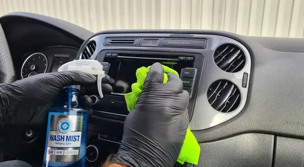 Ręce w czarnych rękawiczkach wykonują polerowanie plastiku na desce rozdzielczej samochodu, używając zielonej ściereczki i płynu.