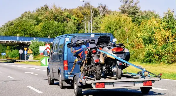 Jak przewieźć motocykl na zwykłej przyczepie? Dwa motocykle bezpiecznie zamocowane pasami na przyczepce za niebieskim vanem.