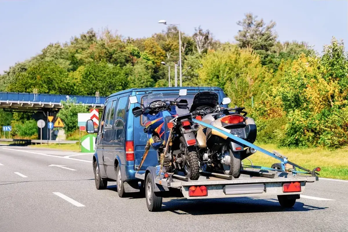Jak przewieźć motocykl na zwykłej przyczepie? Dwa motocykle bezpiecznie zamocowane pasami na przyczepce za niebieskim vanem.