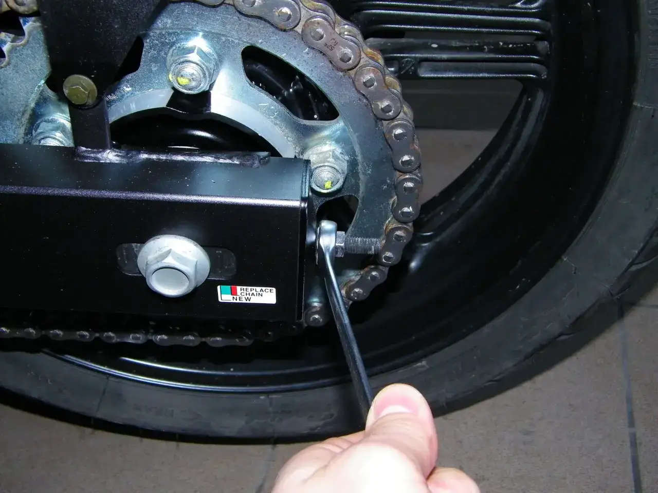 Dłoń dokręca śrubę regulującą naciąg łańcucha w motocyklu. Obok naklejka 