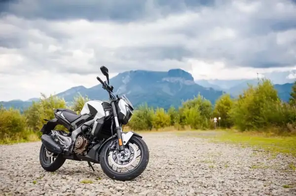 Biało-czarny motocykl Bajaj 400 na kamienistym podjeździe, w tle góry i chmury.