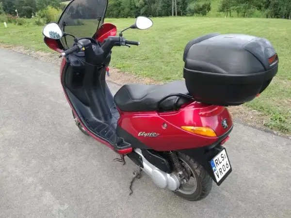 Czerwony skuter Peugeot Elyseo 125 z czarnym kufrem na bagaż, gotowy na podróż po malowniczych drogach.