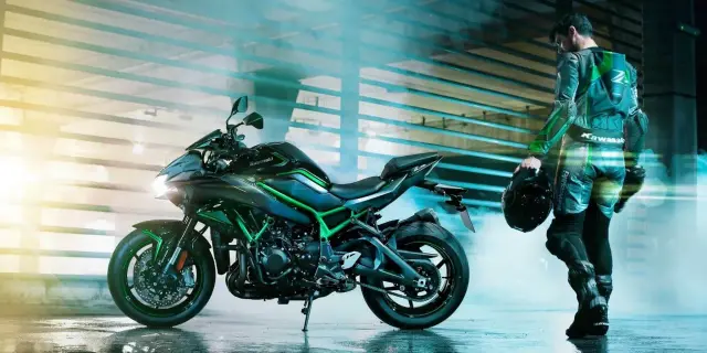 Motocyklista w kombinezonie Kawasaki z kaskiem w dłoni stoi obok czarnego motocykla H2R z zielonymi akcentami.