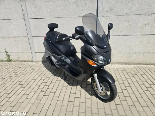 Czarny skuter Piaggio X9 z dużym przednim wiatrochronem, zaparkowany na chodniku z kostki brukowej, obok betonowego muru.